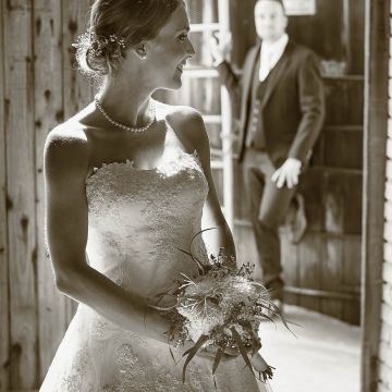 Braut seitlich stehend blickt auf den Bräutigam am Holztor lehnend