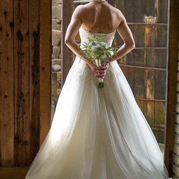 Braut im langen weißen Hochzeitskleid von hinten vor einem Holztor im Gegenlicht
